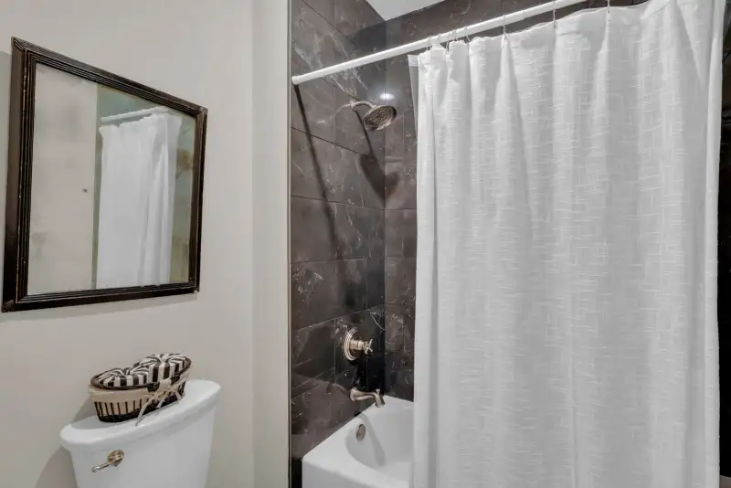 Tub and toilet with dark marble-look tile, a white shower curtain, and a framed mirror on a beige wall.