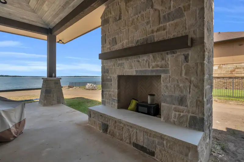 Outdoor stone fireplace on a covered patio, with a lake visible beyond the yard.
