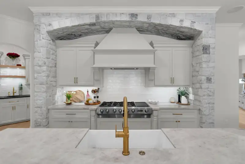 Range set in a stone arch between white cabinets, with a gold faucet and a white farmhouse sink in front.