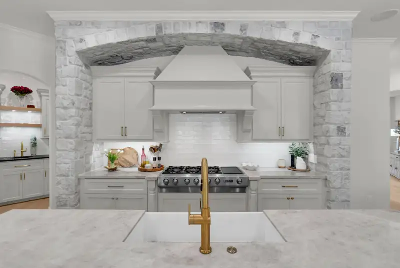 Range set in a stone arch between white cabinets, with a gold faucet and a white farmhouse sink in front.