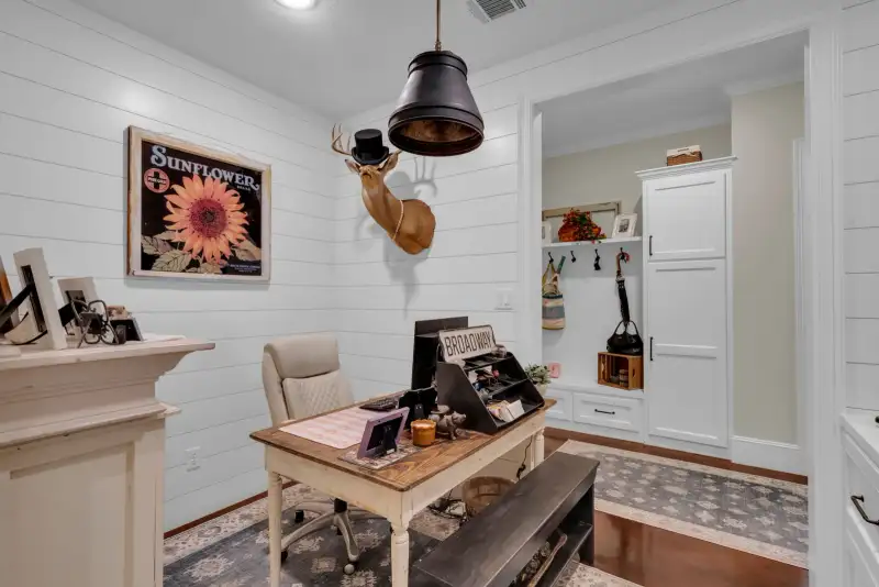 Home office with white shiplap walls, a cream wood desk, a rolling chair, and a framed sunflower print.