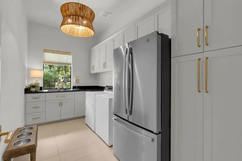Laundry room with a woven pendant light, white cabinets with brass pulls, and a stainless refrigerator.