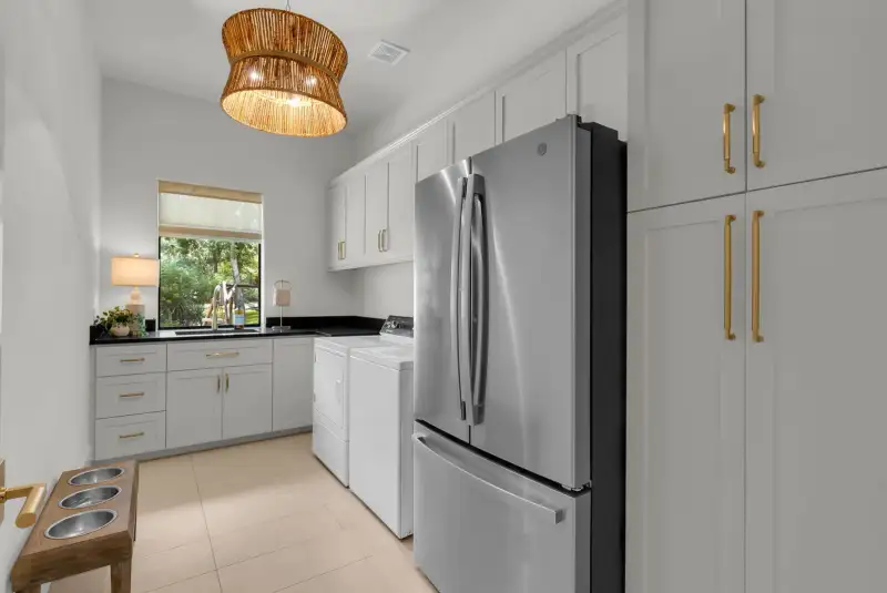 Laundry room with a woven pendant light, white cabinets with brass pulls, and a stainless refrigerator.