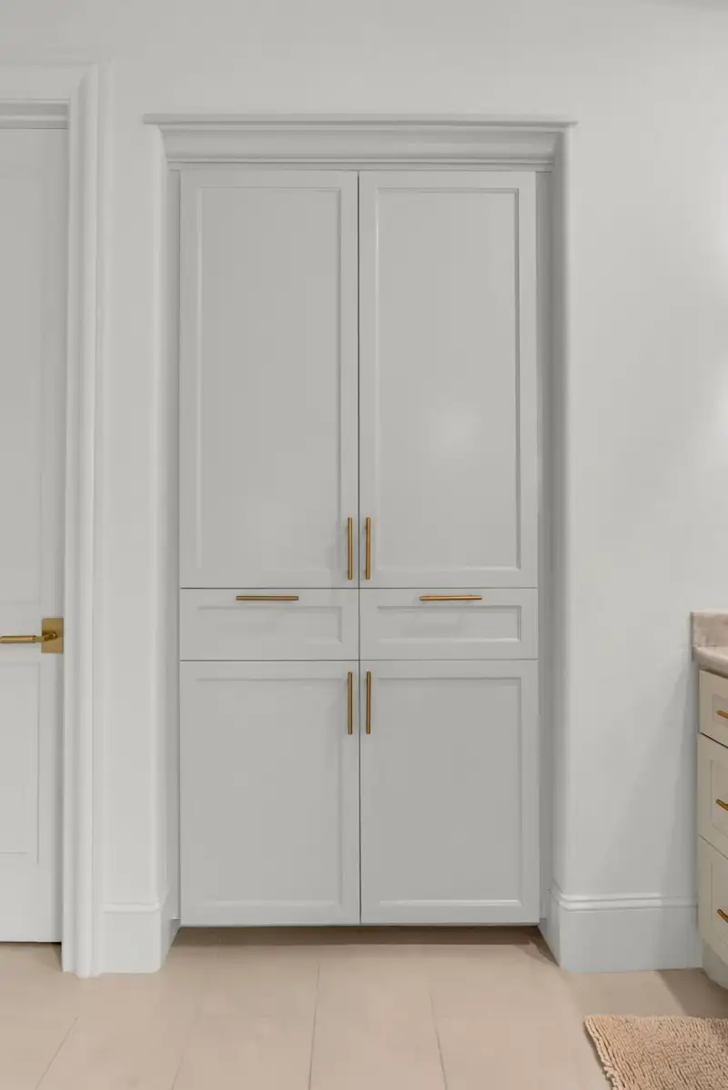 Tall gray storage cabinet with brass pulls and two drawers, beside a door.