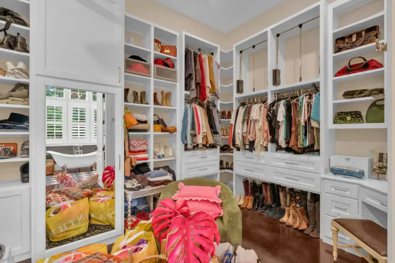 Walk-in closet with white built-in shelving, hanging clothes, a mirrored door, and rows of boots and handbags.