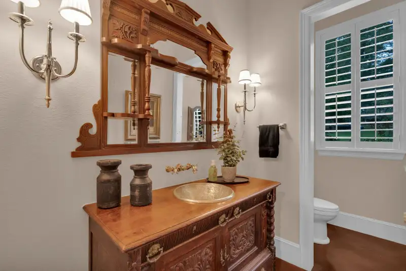 Powder room with a carved wood vanity, a gold vessel sink, wall sconces, and plantation shutters.