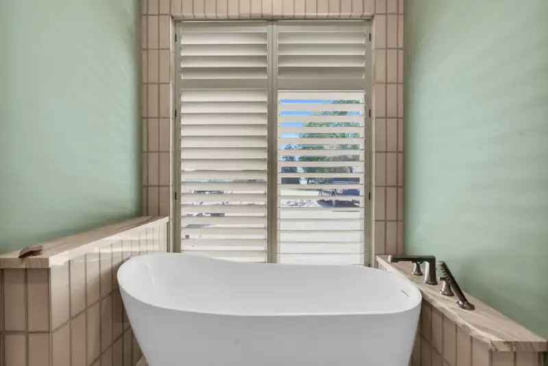 Freestanding white tub in front of a shuttered window, with tile around the base.