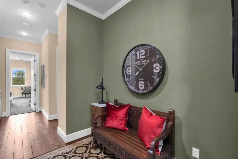 Wood bench with red pillows on a patterned rug, beneath a wall clock on a green wall.
