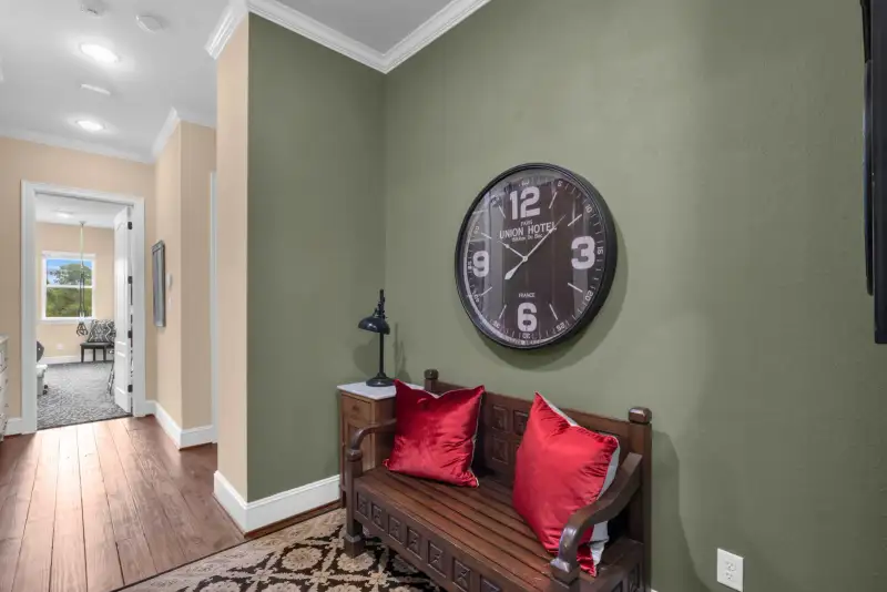 Wood bench with red pillows on a patterned rug, beneath a wall clock on a green wall.