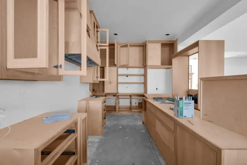 Narrow kitchen with unfinished wood cabinets and drawers, and a window with trees at the end.