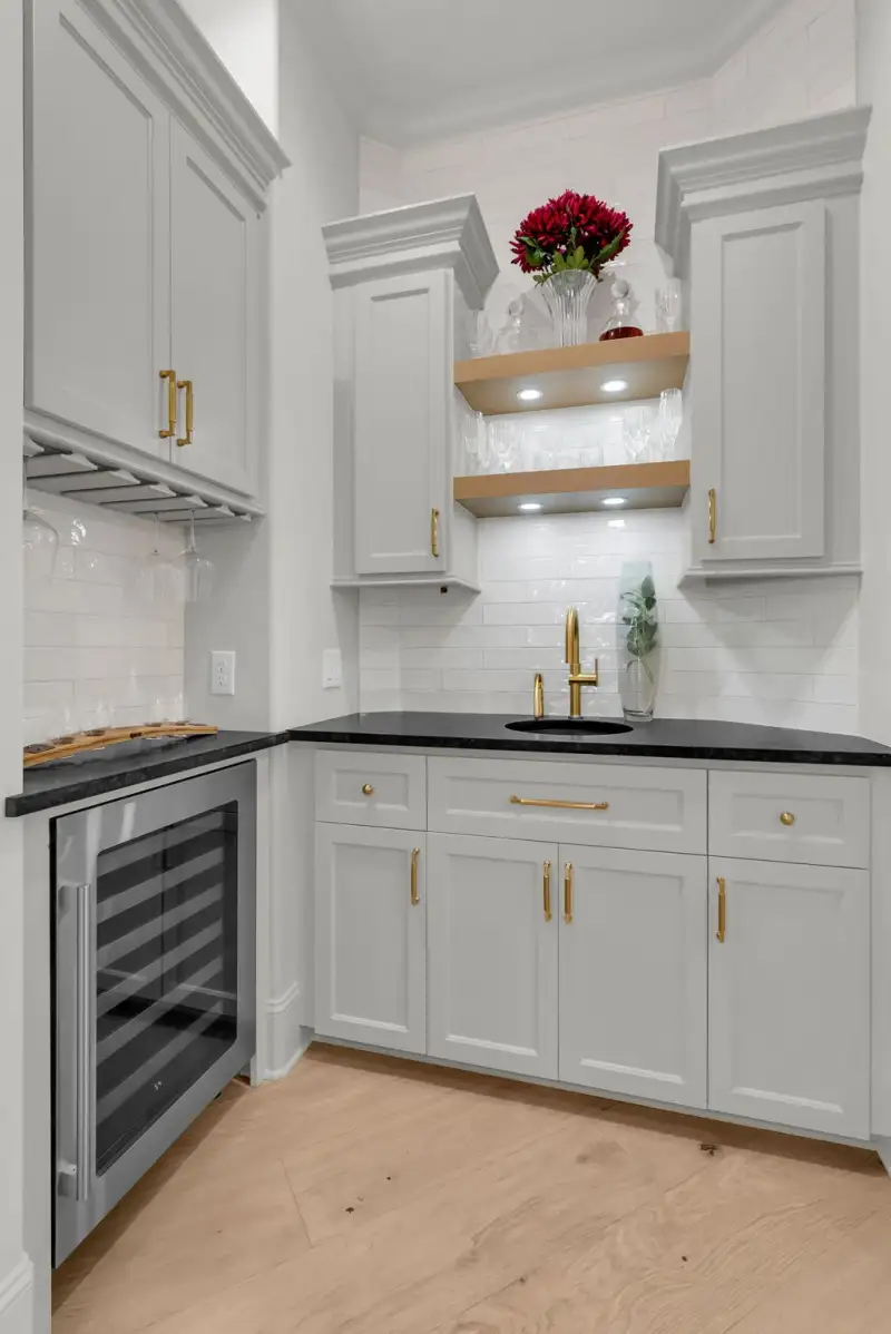 Bar area with a wine refrigerator, open wood shelves, dark countertops, and a gold faucet over a sink.