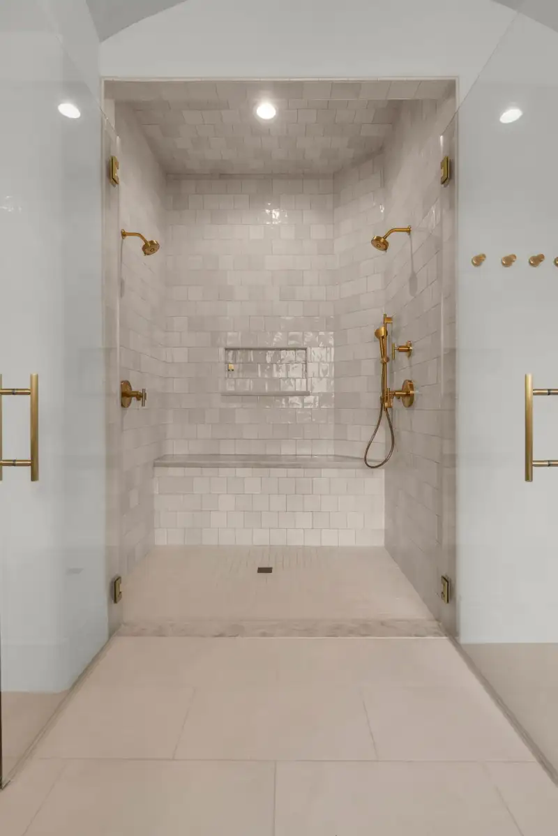 Walk-in shower with tile walls, a built-in bench, two brass shower heads, and a recessed niche.