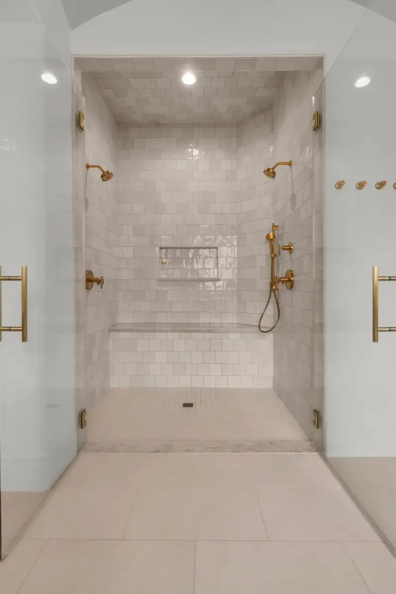 Walk-in shower with tile walls, a built-in bench, two brass shower heads, and a recessed niche.