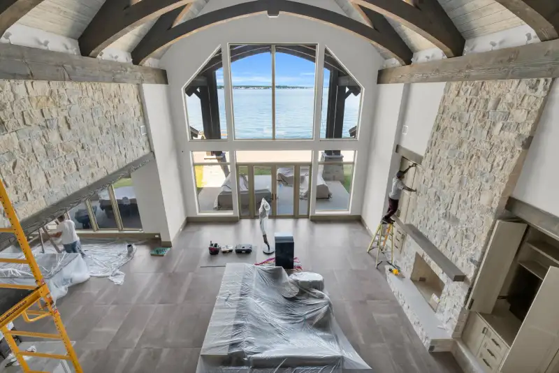 Two-story room with tall windows, a stone fireplace, and workers on site.