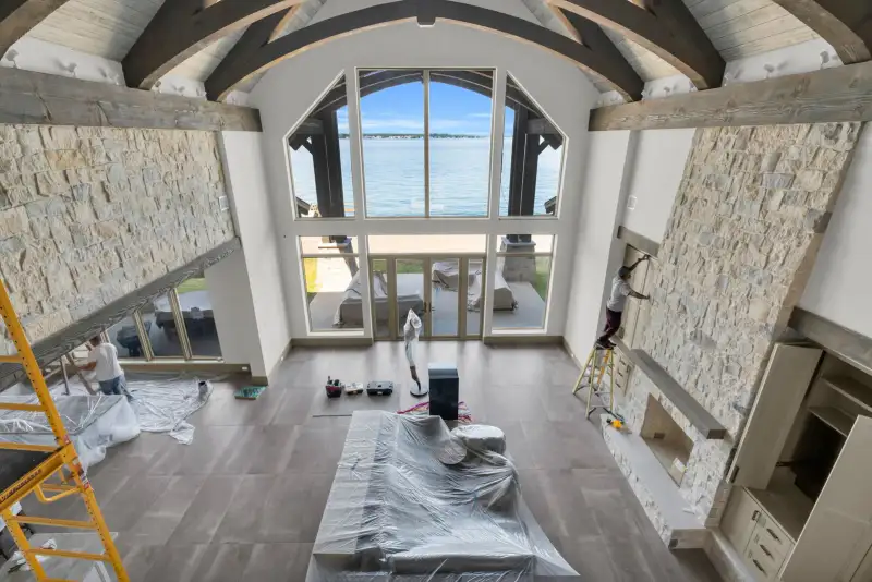 Two-story room with tall windows, a stone fireplace, and workers on site.