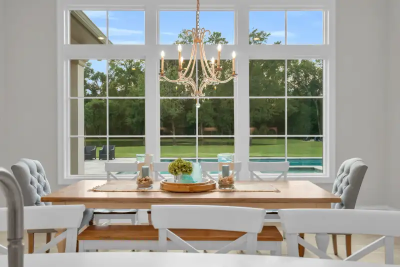Breakfast table with a wood top, white crossback chairs, and a large window wall overlooking a pool.
