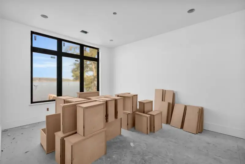 Unfinished room with a black-framed window and cardboard cabinet boxes stacked on the concrete floor.