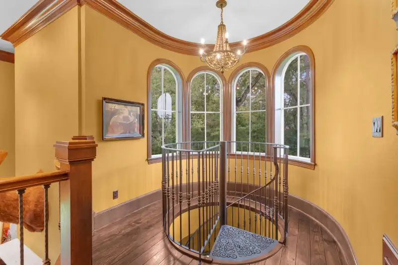 Spiral iron staircase with a patterned landing rug beneath arched windows and yellow walls.