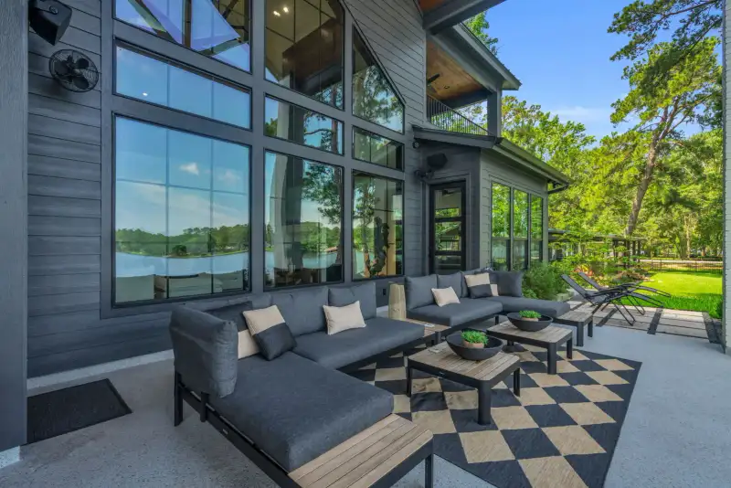 Outdoor living area with gray sectional seating, a checkered rug, and tall windows overlooking trees.
