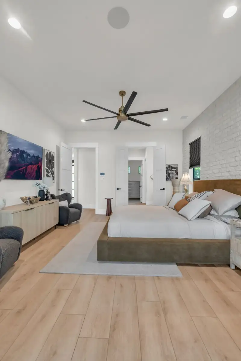 Bedroom with light oak flooring, a brown upholstered bed, and a white brick accent wall beside windows.