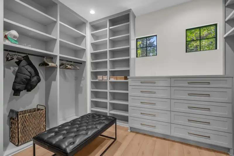 Walk-in closet with white shelving, gray drawers, a tufted leather bench, and two black-framed windows.
