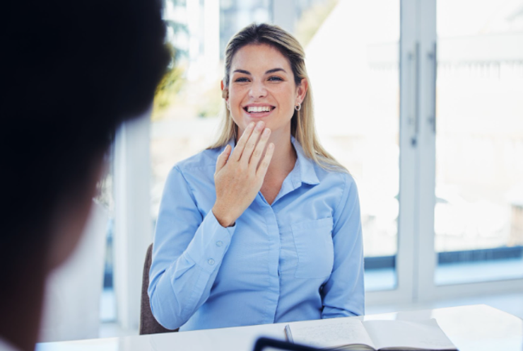 Deaf employee saying Thank you to a colleague in sign language. She is wearing a blue blouse and sitting at a white desk with a notebook open in front of her.
