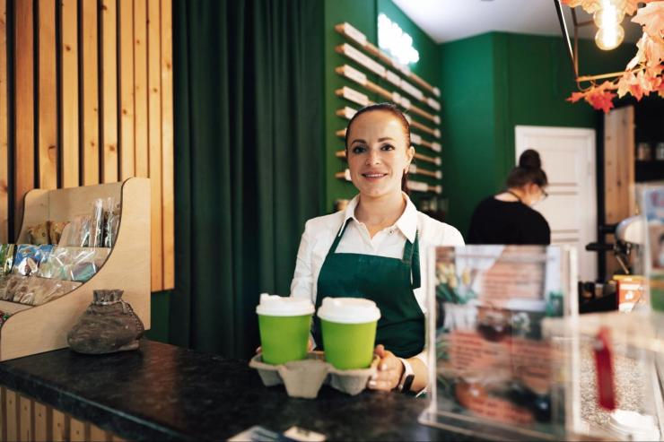 A young barista woman wearing a green apron is holding a tray of coffee.