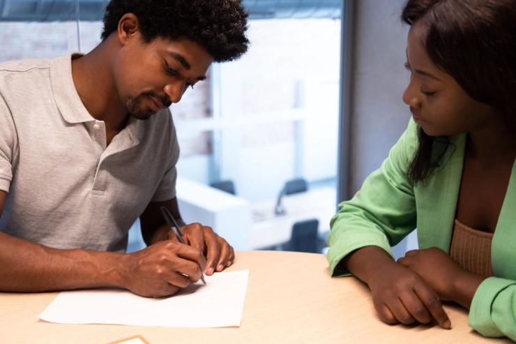 A black man is writing on paper before a seated black woman. 