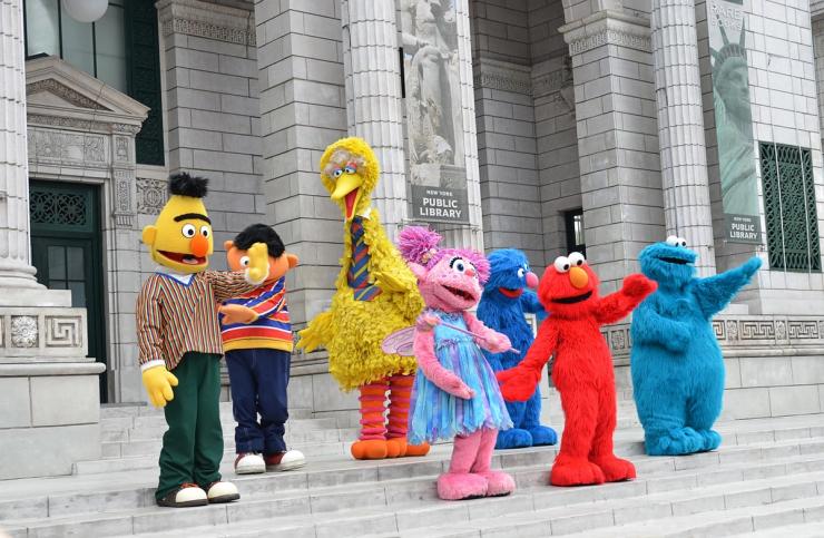 The Sesame Street cast is standing on steps in front of a library. 