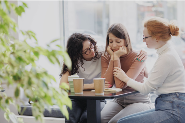 Three young women sitting at a table with coffee cups. Two women comfort the third woman, who seems upset, with her hands on her face. Photo by myjuly.