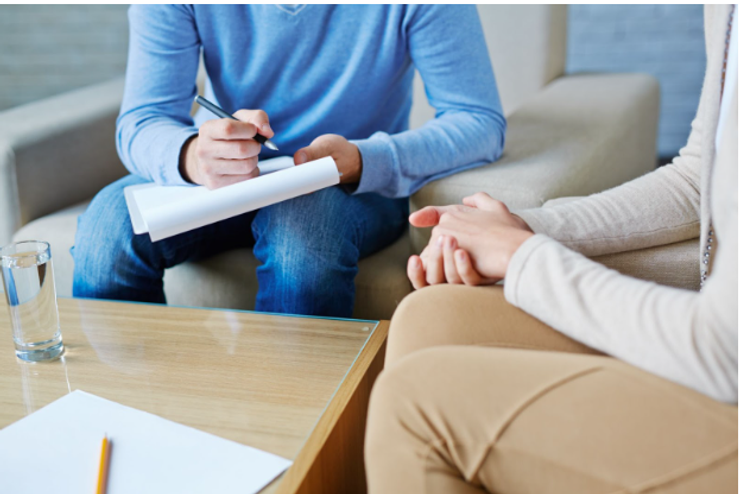 A woman patient sitting with hands together. She is talking with a male counselor who is writing on a notepad. Their faces are hidden. Photo by Pressmaster. 