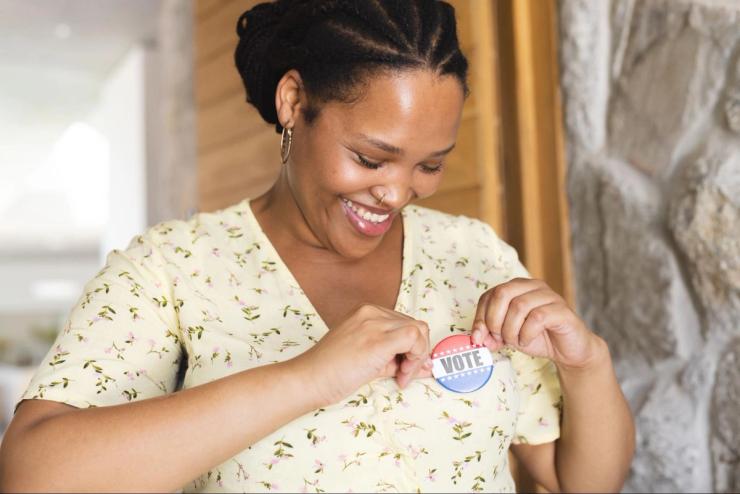 A young black woman affixes a button to her shirt that says “VOTE.” 