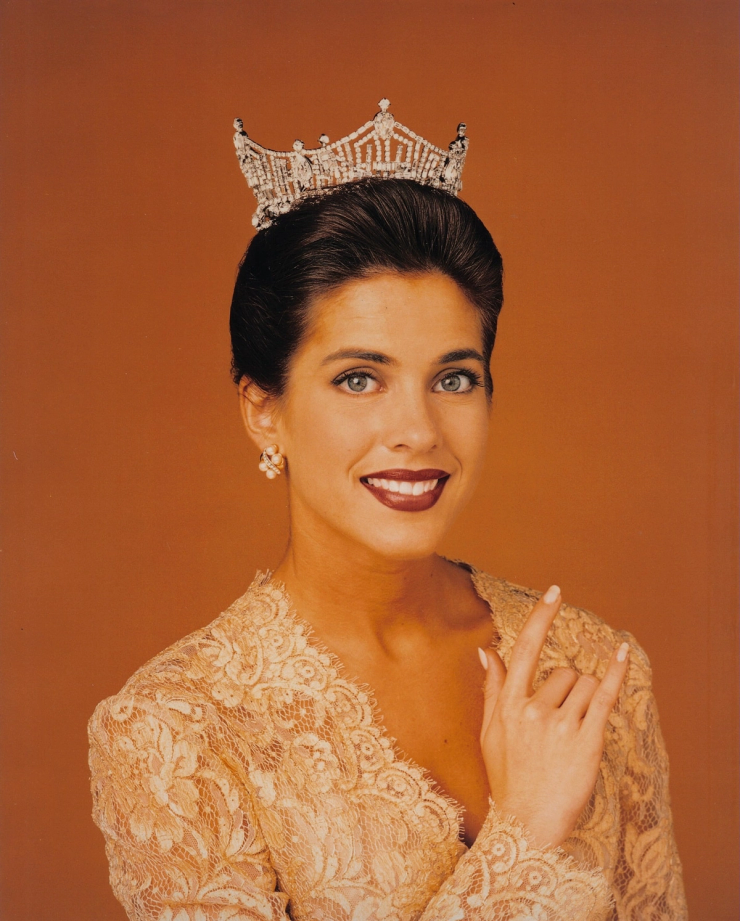 woman wearing a crown and lace dress posing for a formal portrait against an orange background