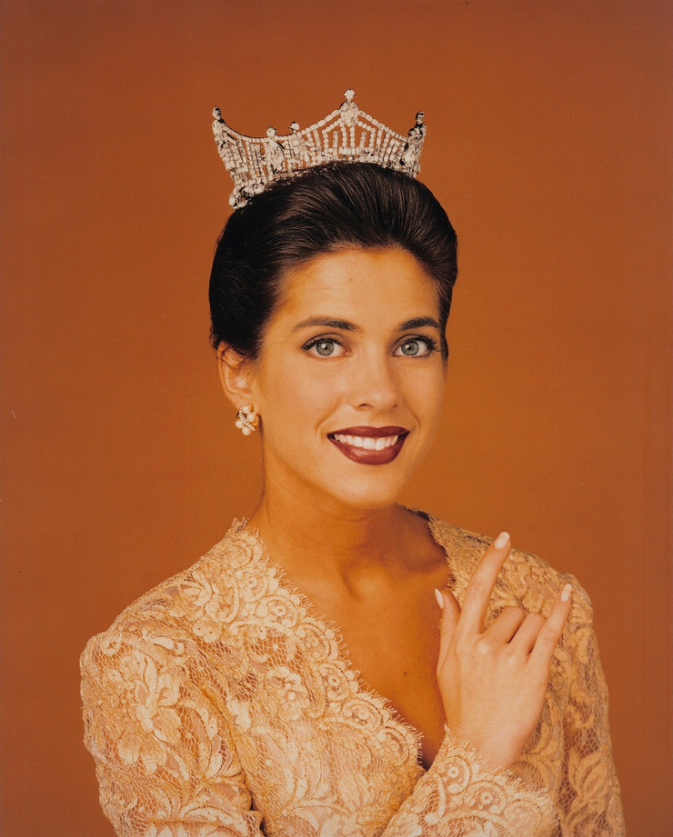 woman wearing a crown and lace dress posing for a formal portrait against an orange background