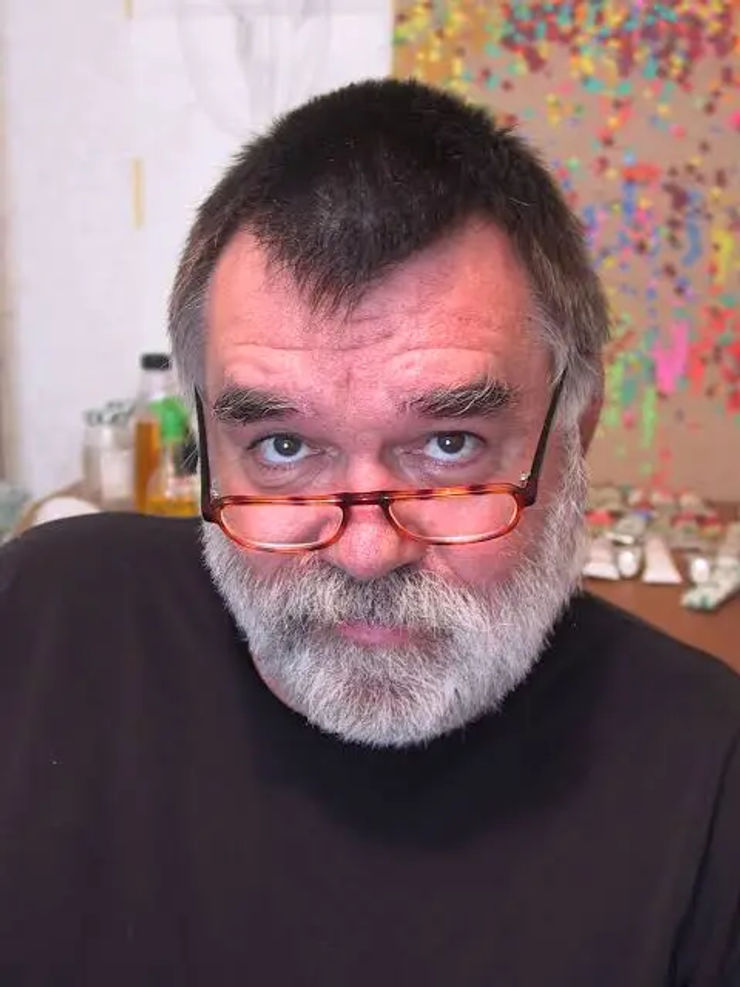 Middle-aged man with a gray beard wearing glasses looks directly at the camera in an indoor setting with a colorful background