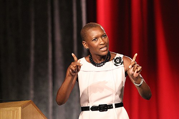 Woman speaking at a podium gesturing with both hands on stage with a red curtain backdrop during a presentation or speech
