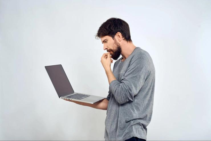 A bearded white man with dark hair and a gray shirt is holding a laptop computer and thinking. 