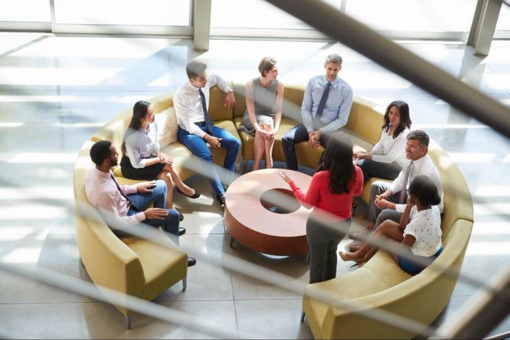 A group of businesspeople sitting on a round couch in front of large windows.