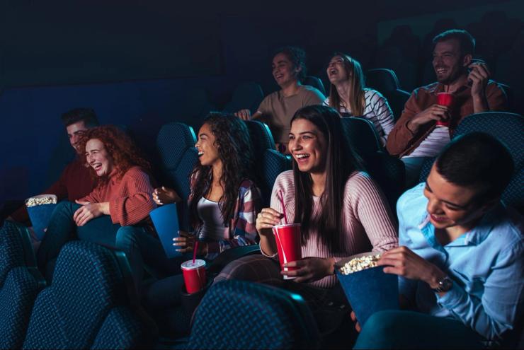 A group of people laughing in a movie theater while drinking and eating popcorn.