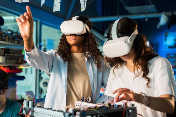 Two women are wearing Virtual Reality headsets while standing.