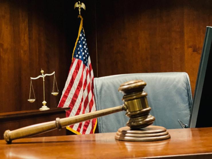 A gavel rests ahead of an American flag and weighted scales on a judge's desk.