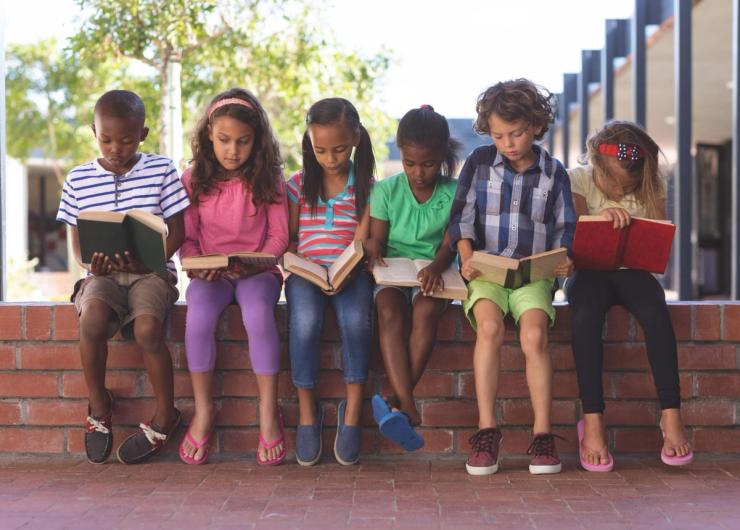 A group of young children sitting on a brick wall and reading books.