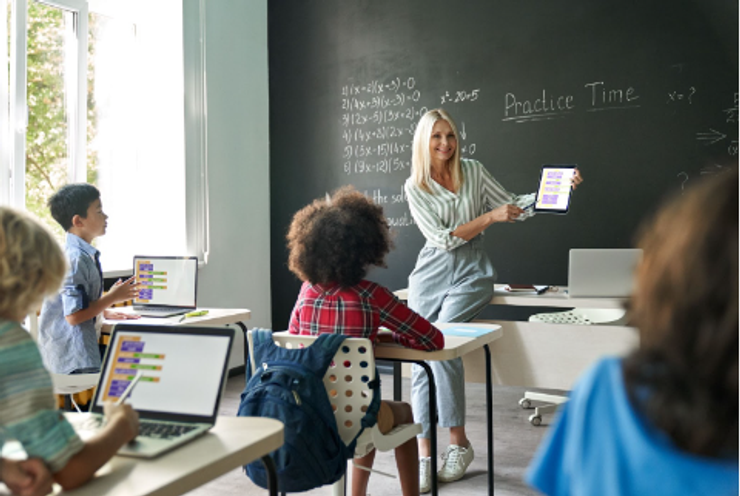 Smiling teacher sitting on a desk while holding iPad and giving a math lesson to students