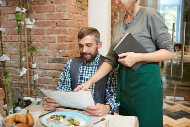 A waiter pointing at a menu in front of a man sitting down. 