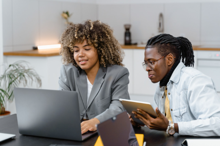 Two colleagues sitting next to each other. One is using a laptop and the other is holding an iPad. Photo by MART PRODUCTION. Pexels