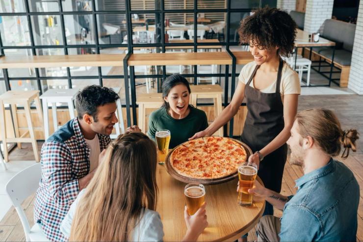 A group of people sits around a table as a waiter serves a pizza.
