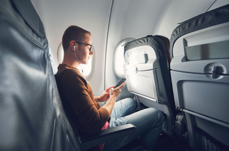 Young male traveler on airline flight using a smartphone connected to in-ear headphones