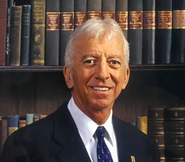 Older man in a suit and tie smiling in front of a bookshelf filled with hardcover books in an office or library setting