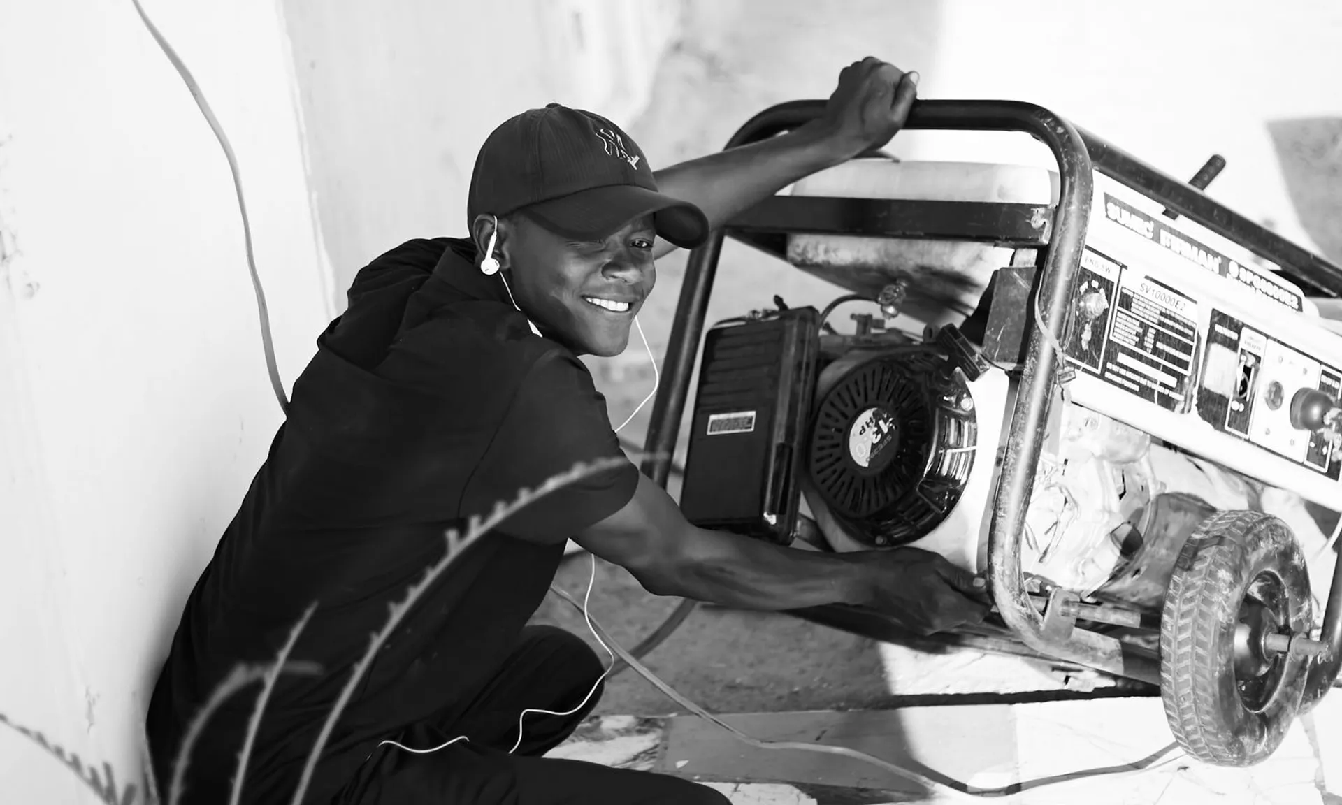 A cheerful young man wearing a cap and earphones operating a portable generator outside.