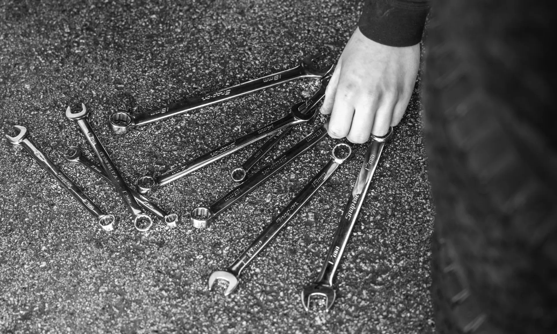 Black and white image of wrenches spread on asphalt with a hand picking up one.